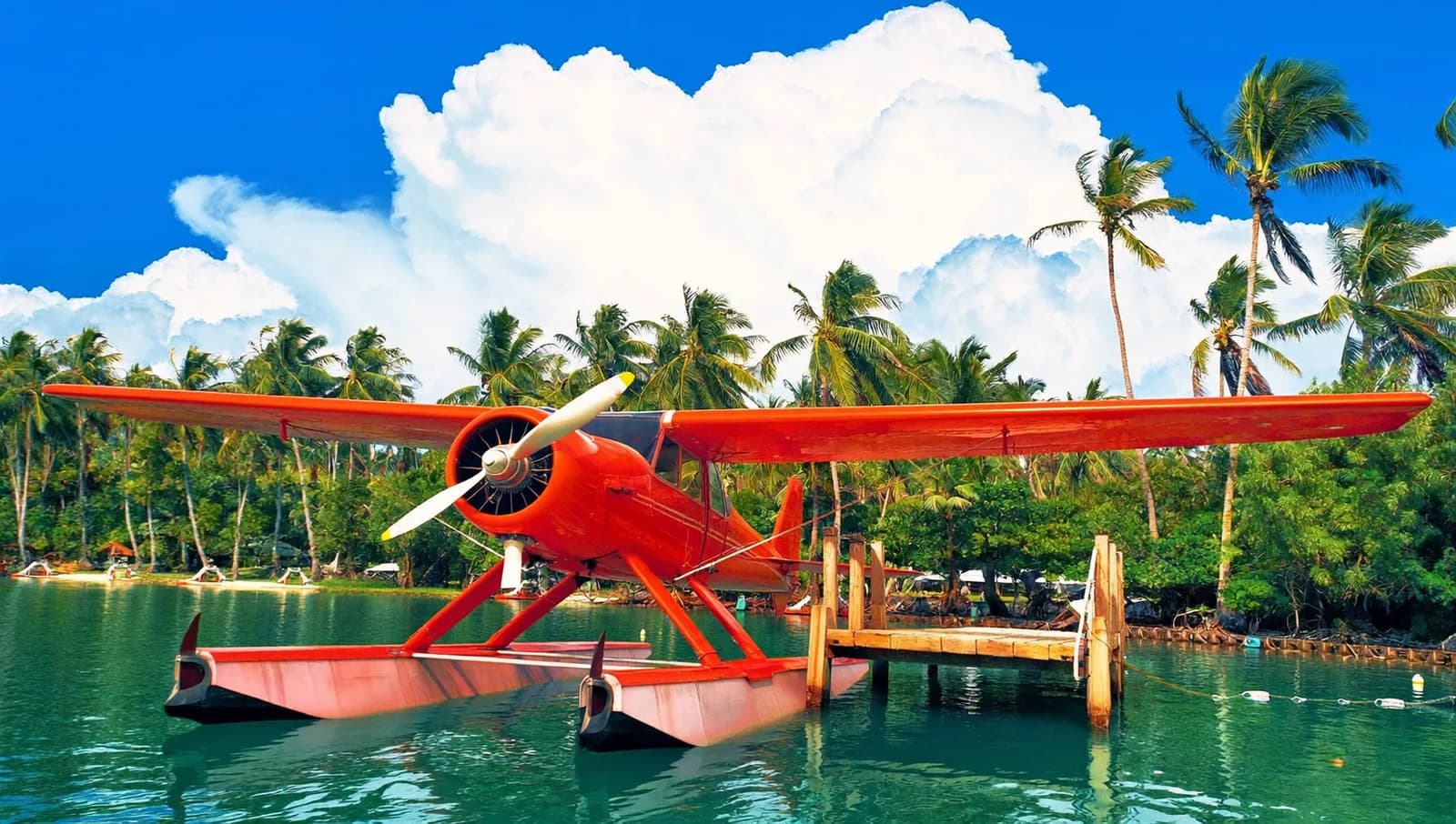 A red vintage seaplane floating on a turquoise tropical lagoon beside a small wooden dock, palm trees swaying, towering white cumulus clouds, bright midday sun, vivid travel photography