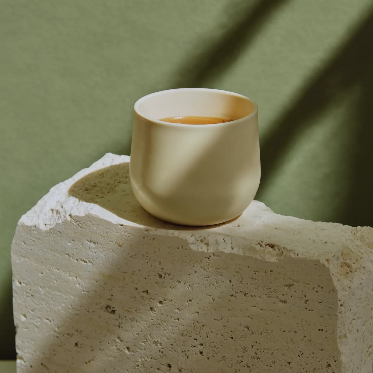 Minimal product still life of an unbranded matte ceramic coffee cup on a travertine block, sharp diagonal shadow from a window, sage green backdrop, soft morning light, premium studio photography