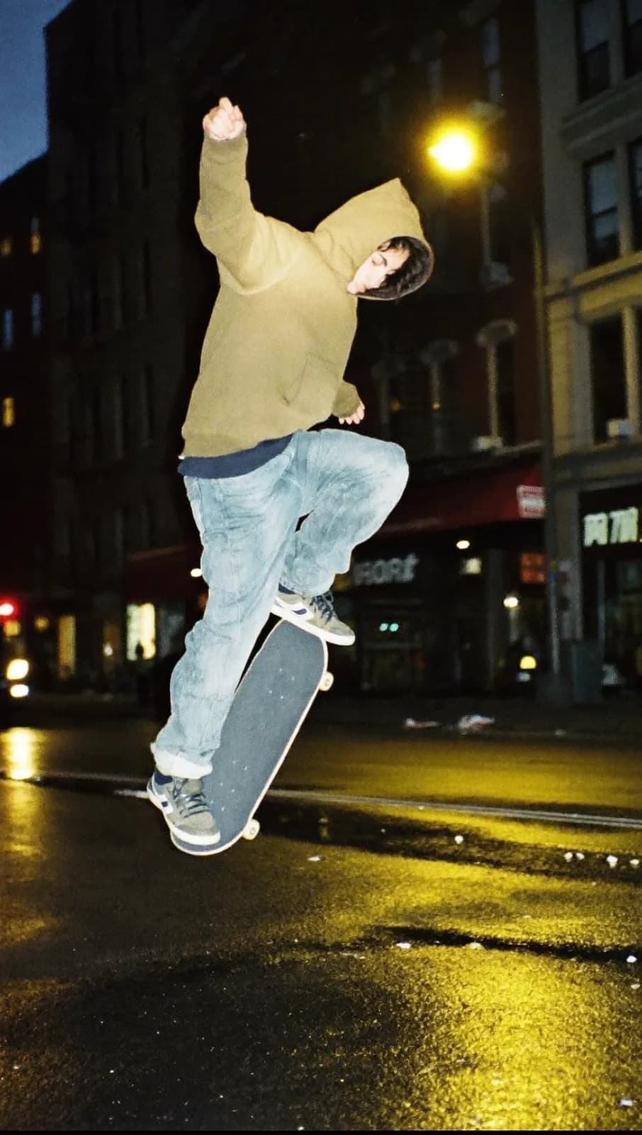 Candid street photo of a skateboarder mid-kickflip under a flickering sodium streetlight at night, motion blur, wet asphalt reflections, gritty 35mm film look, vertical frame