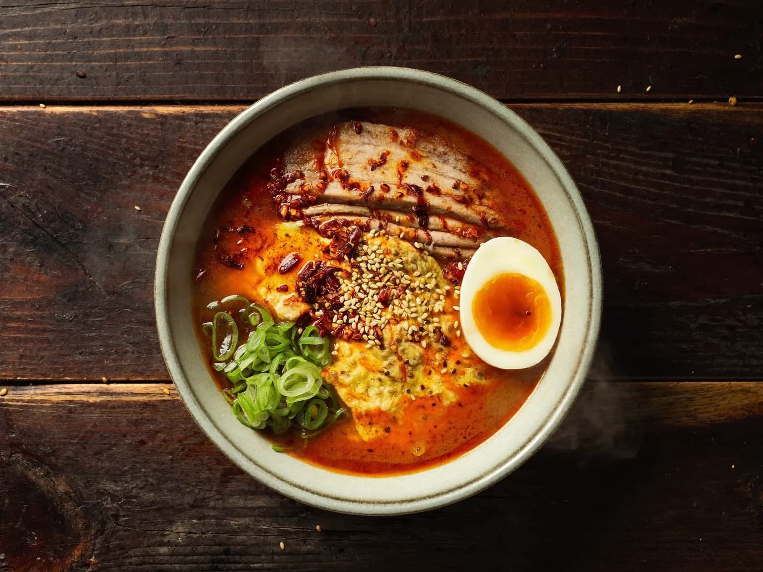 Overhead food photography of a steaming bowl of spicy ramen with a jammy soft-boiled egg, scallions, chili oil swirls and sesame seeds on a dark wooden table, moody side light, rich textures