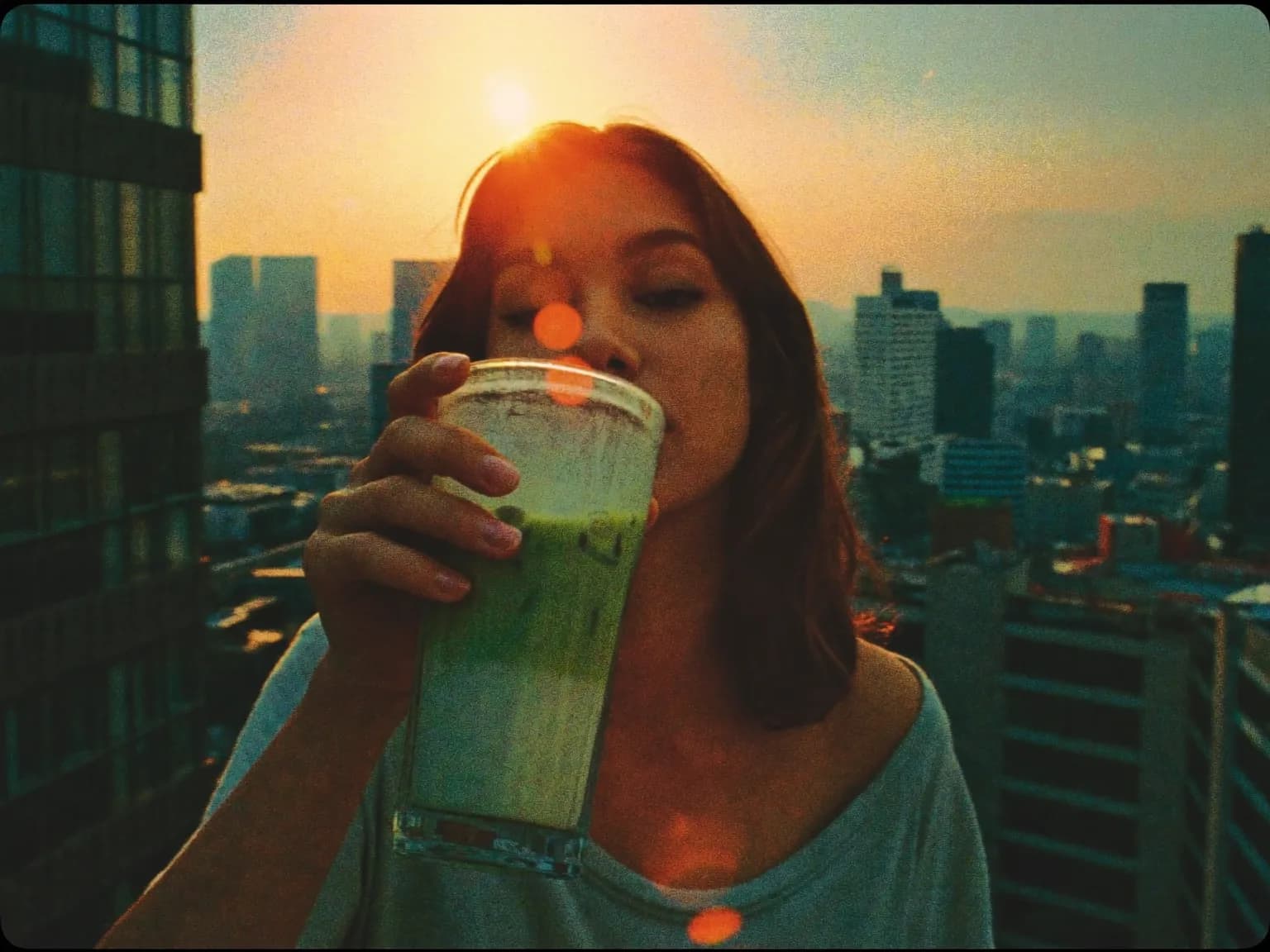 Film still, a young woman holding a tall glass of iced matcha latte on a city rooftop at golden hour, 35mm anamorphic lens, low-angle medium shot, warm backlight with lens flare, teal-and-orange color grade, hazy atmosphere, shallow depth of field, film grain