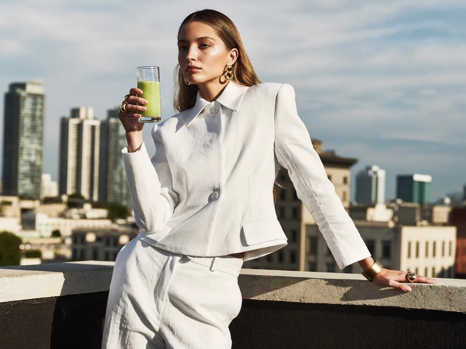 High-fashion editorial photograph, a young woman holding a tall glass of iced matcha latte on a city rooftop at golden hour, wearing an architectural white linen suit and gold jewelry, poised model pose, crisp hard sunlight, clean magazine composition with bold negative space